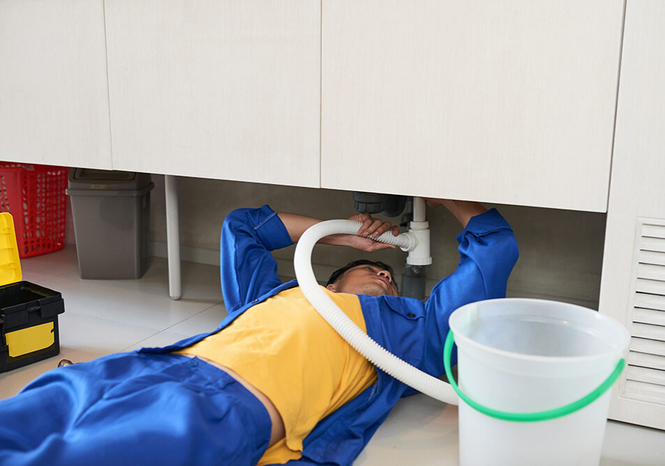 Plumber in uniform lying on the floor and fixing plastic sink pipe in kitchen