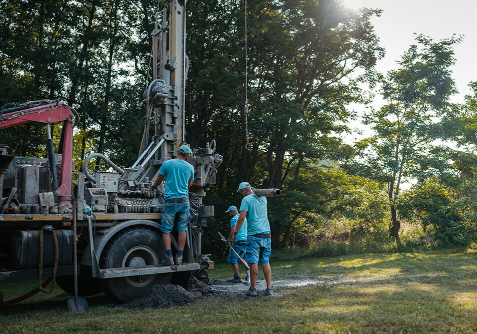 Water well drilling rig, truck preparing to boring dowin into the earth. Professional workers operating drilling machine, shoveling rubble. A rotary drill rig using bits to bore into ground and loosing the soil and rocks. Mud and muddy water is spitting out when drilling borewell. Banner with copy space.